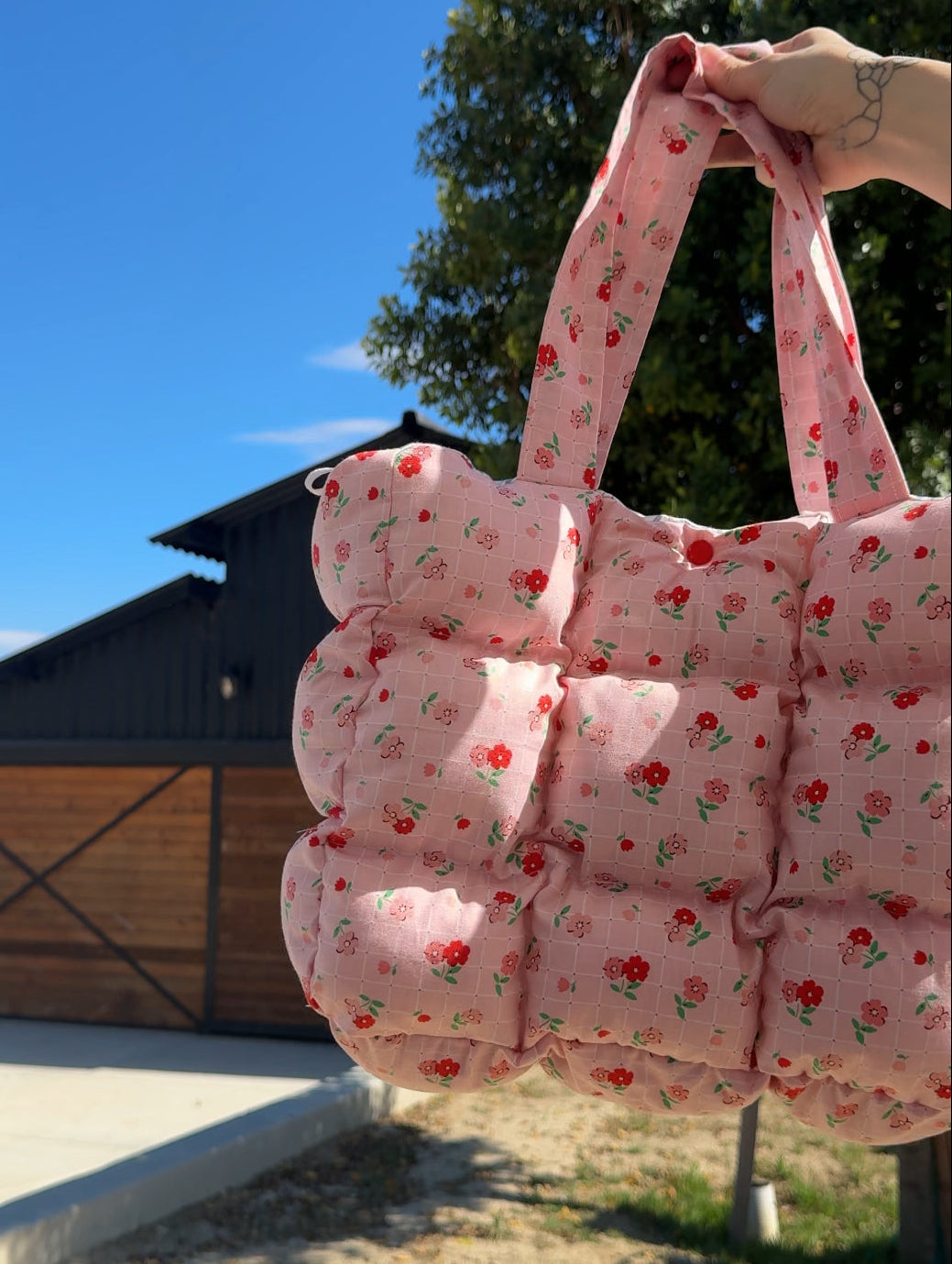 Pink floral quilted bag held outdoors with a clear blue sky and wooden building in the background