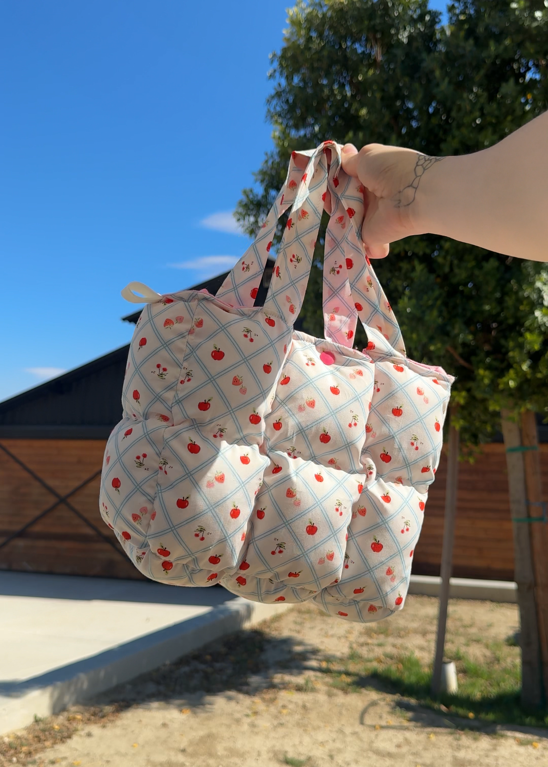 Person holding a patterned reusable bag outdoors with a clear blue sky and trees in the background.