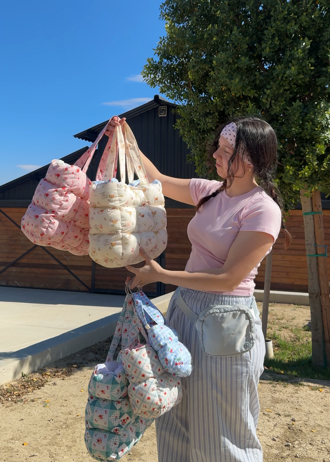Woman holding multiple puffy quilted bags outdoors with a clear blue sky and trees in the background
