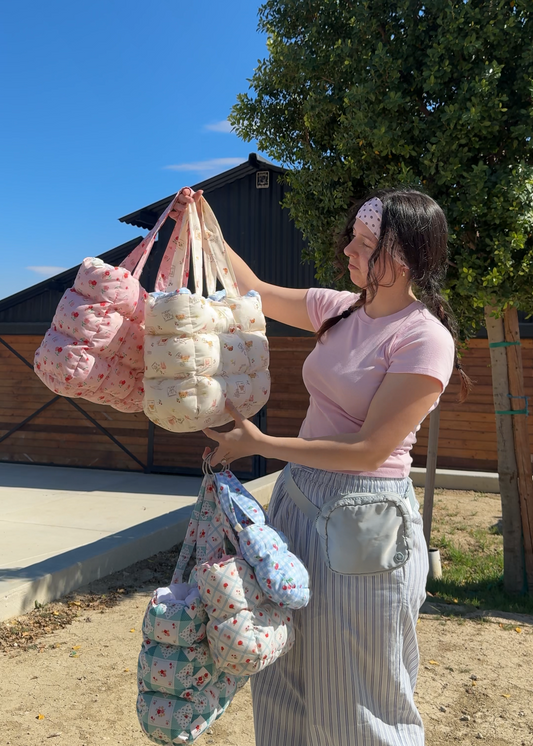 Woman holding multiple puffy quilted bags outdoors with a clear blue sky and trees in the background
